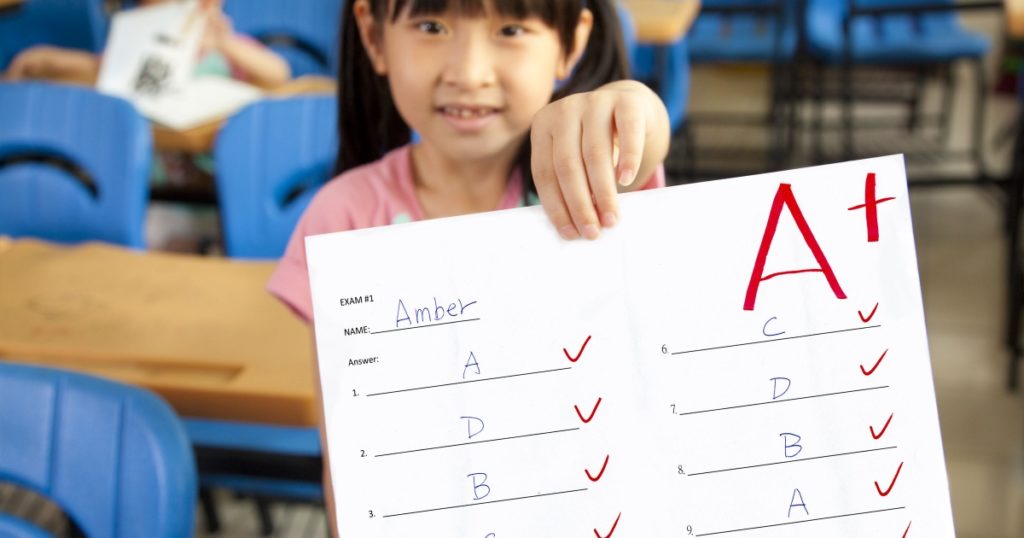 smiling little girl showing exam paper with a plus in the classroom