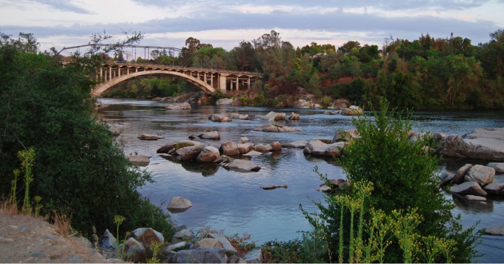 Rainbow Bridge in Folsom California in the early morning with green trees along the river banks and big boulders reflecting in the water