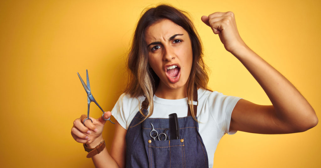 Young beautiful hairdresser woman holding scissors over yellow isolated background annoyed and frustrated shouting with anger, crazy and yelling with raised hand, anger concept
