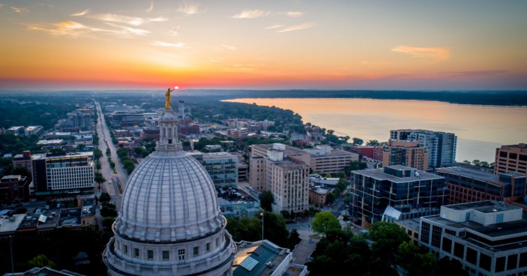 Wide aerial shot of a dramatic, fiery sunrise over the Wisconsin State Capitol. Statue atop the dome appears to hold the rising sun.