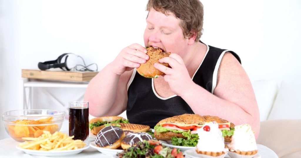 Fat man eating a lot of unhealthy food, on home interior background