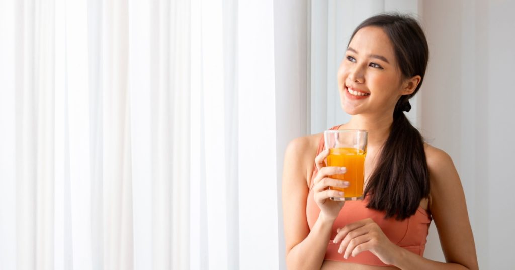 Healthy asian woman drinking a glass of orange juice for refreshment while wearing sportswear for nutrition and vitamin c supplements at the window