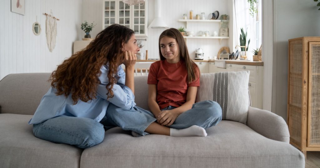 Teenage daughter sharing secrets with young loving supportive mother, parent mom talking chatting with adolescent girl while sitting together on sofa at home. Healthy parent-teen relationships