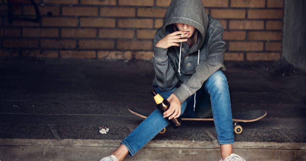 Teen girl in hood holding a beer bottle and smoking the cigarette on the steps in the alley