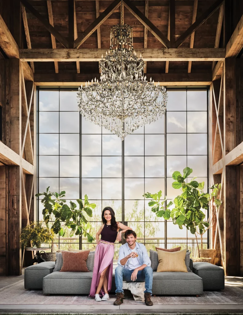 Mila Kunis and Ashton Kutcher pose in front of a big window. Trees in the background.