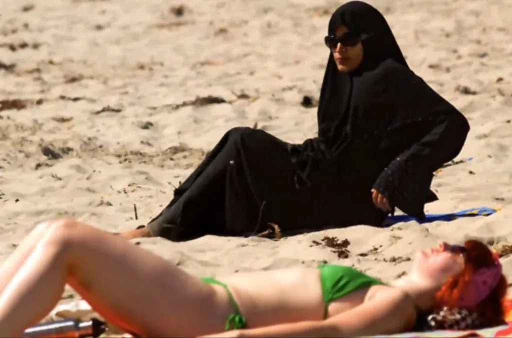 Here, a woman in a bikini lounges on a beach adjacent to another woman clad in a burqa