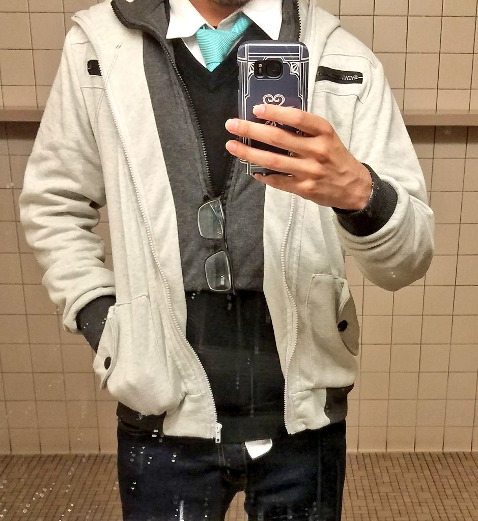 Person wearing layers and taking a picture on a cell phone in a mirror. Tiles on the wall and floor