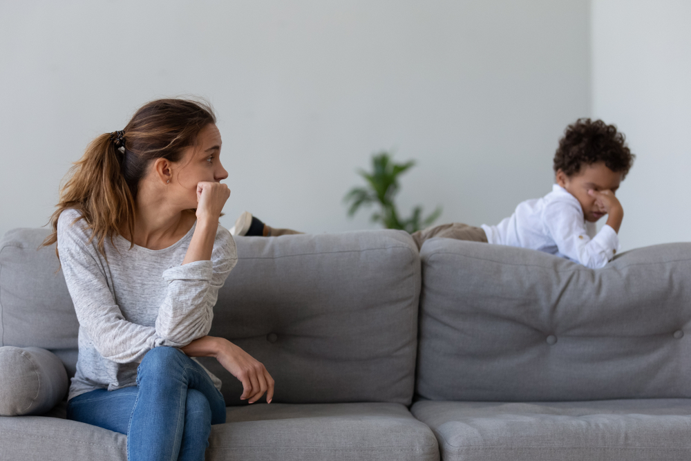 Unhappy young woman sitting on sofa in living room at home, looking at stressed offended punished upset little kid son, lying on couch. Sad mommy feeling guilty about quarrel conflict with child.
