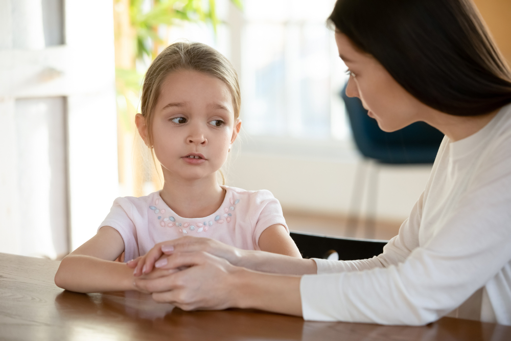 Head shot unhappy small child girl sitting at table with worrying mother, sharing school problems. Compassionate caring attentive mommy having trustful conversation with unhappy offended daughter.
