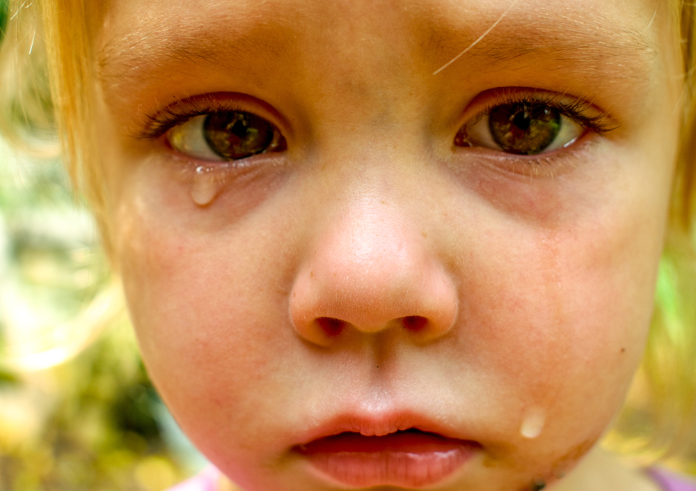 A close-up of a sad little girl with brown eyes crying with tears rolling down her face looking strait into your eyes- A little girl crying with tears in her eyes
