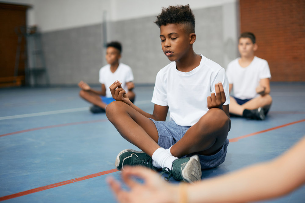 African American schoolboy and his friends meditating in lotus position during PE class at school gym.
