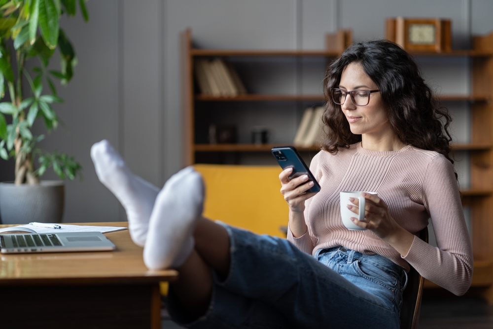 Woman freelance writer rest or procrastinate at workplace. Businesswoman in eyewear distracted from work on laptop scrolling social media on smartphone. Freelancer holding mobile phone and browsing