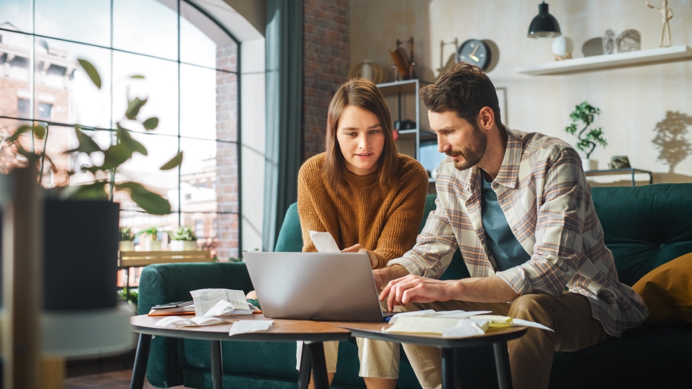Stressful Accounting at Home: Couple Using Laptop Computer, Sitting on Sofa in Apartment. Young Family Filling Tax Forms, Mortgage Documents, Bills, Checks, Balances, Invoices are in Order