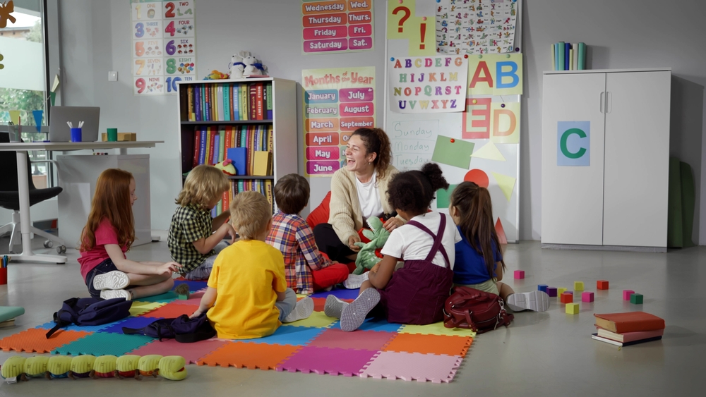 children sitting on floor while caring teacher explains lesson using toy in kindergarten. Elementary school students and teacher sit in circle in classroom
