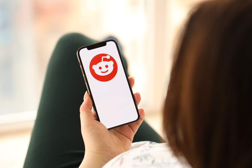 KYIV, UKRAINE - APRIL 1, 2024 Reddit icon on smartphone screen in young female hands indoors. Woman using iPhone app at home