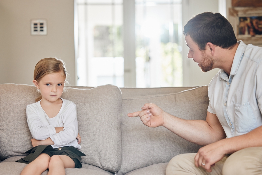 Upset, child and dad with discipline on sofa for bad behaviour, confrontation or punishment in living room. Home, disappointed and strict man pointing at young girl for fight, mistake or scolding
