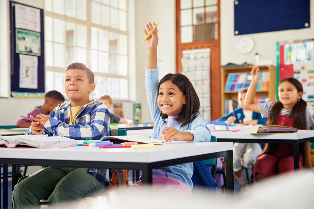 Excited hispanic little girl raising hand in elementary classroom. School children during the lesson trying to answer question in primary school. Cute little latin girl raising hand at school.
