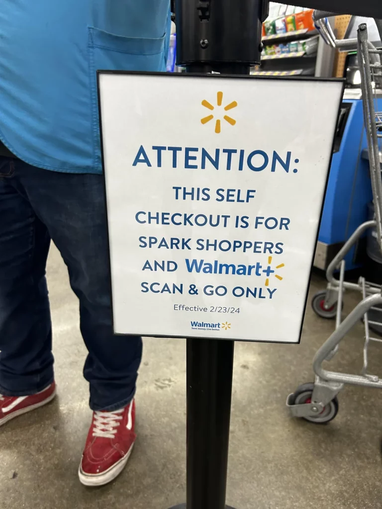 Walmart self-checkout sign with a cart, red shoes, and a blue t-shirt in the background. 