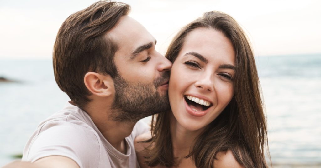 Image of young happy man kissing and hugging beautiful woman while taking selfie photo on sunny beach