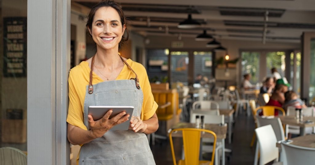 Portrait of happy woman standing at doorway of her store. Cheerful mature waitress waiting for clients at coffee shop. Successful small business owner in casual wearing grey apron standing at entrance