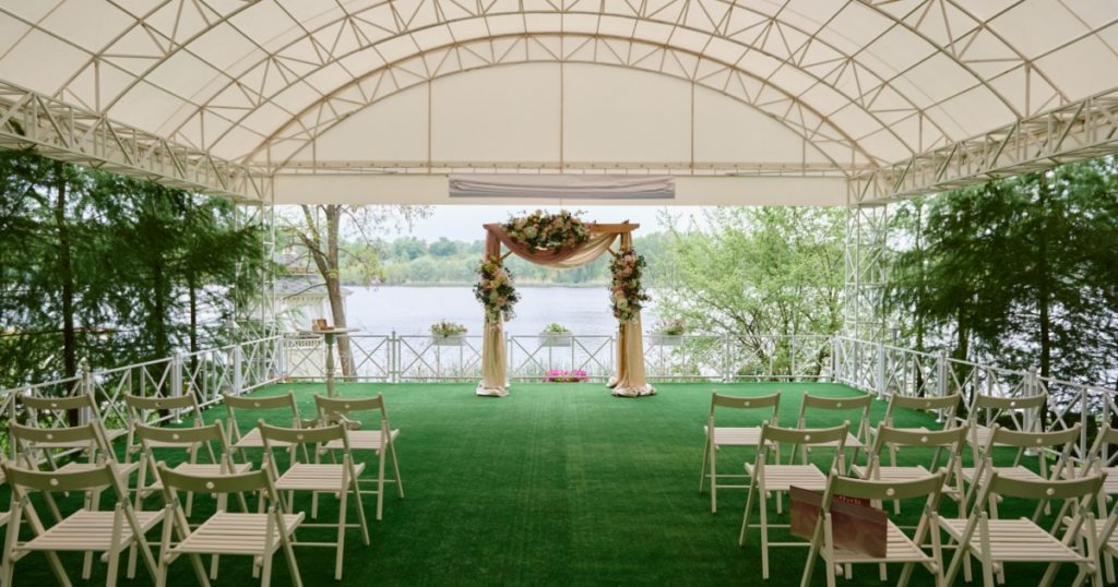 Place for wedding ceremony in tent outdoors, copy space. Wedding arch decorated with flowers and chairs on each side of archway outdoors. Wedding setting