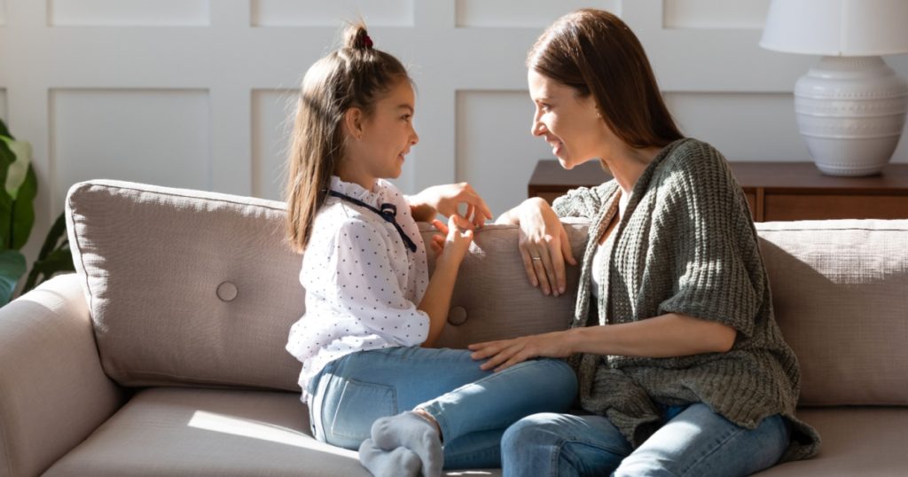 Caring mother and little daughter chatting, sitting on cozy couch in living room, pretty girl sharing secrets with loving mum, having pleasant conversation, good trusted family relationship