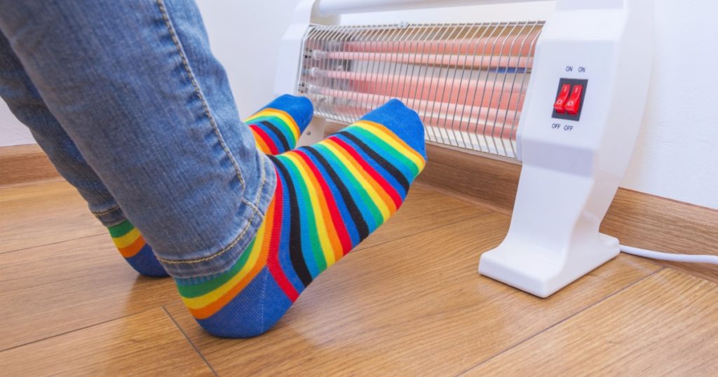 A woman wearing bright rainbow-colored socks warms their frozen feet near a home electric heater. Infrared halogen heater at home.