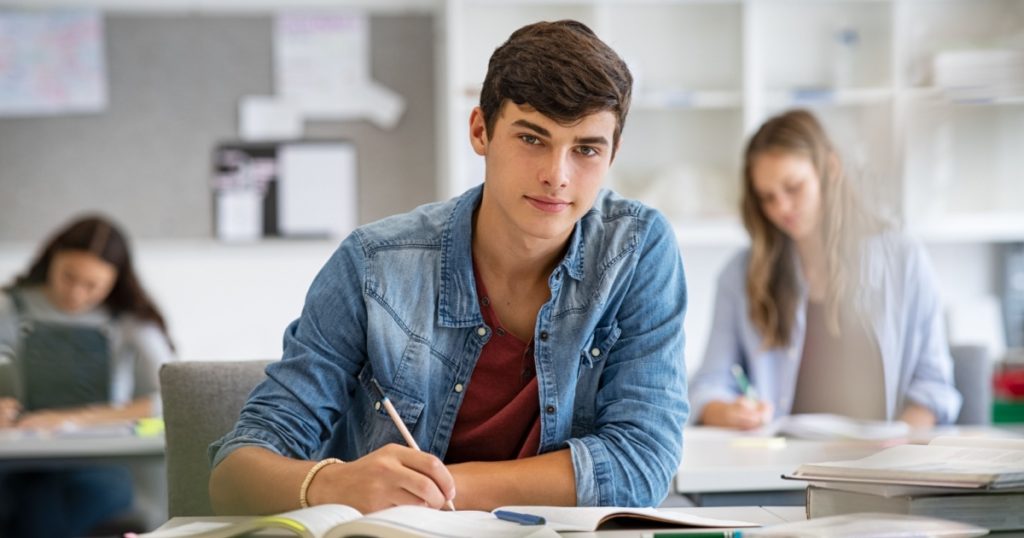 Happy student taking notes while studying in high school. Satisfied young man looking at camera while sitting at desk in classroom. Portrait of college guy writing while completing assignment.