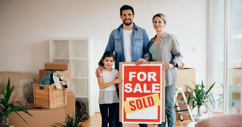 Happy family standing in their new home while holding sold real estate sing and looking at camera.