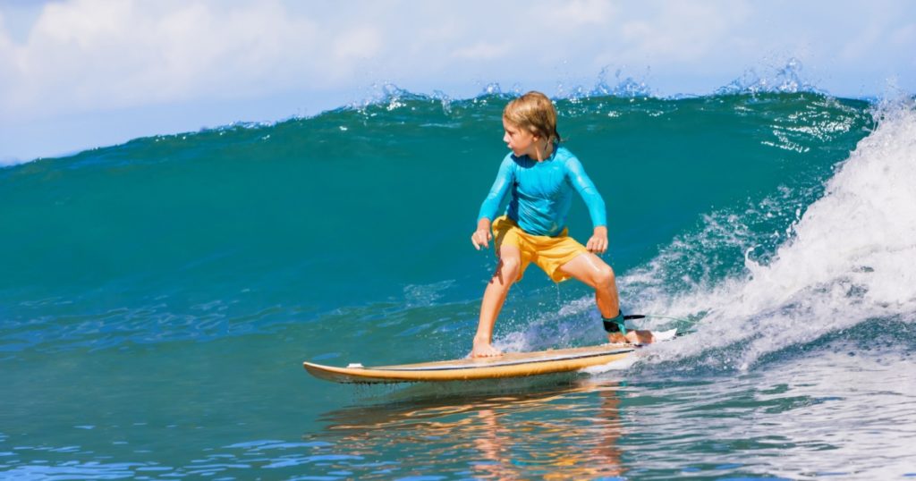 Happy surf boy - young surfer learn to ride on surfboard with fun on sea waves. Active family lifestyle, kids outdoor water sport lessons, swimming activity in surfing camp. Summer vacation with child