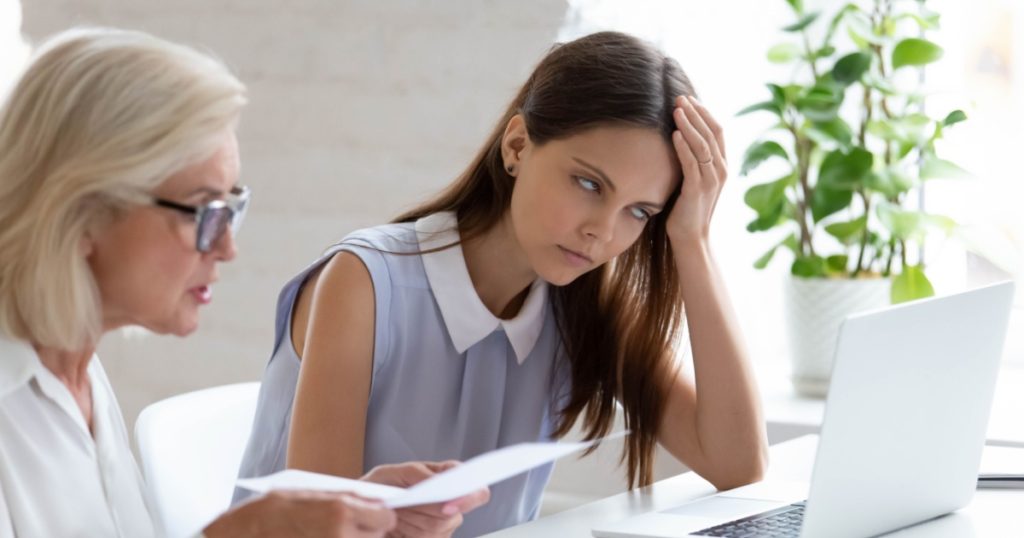 Unhappy young female employee bored with middle-aged colleague discuss paper document at meeting. Tired woman worker bothered annoyed by senior coworker lecture about paperwork at team
