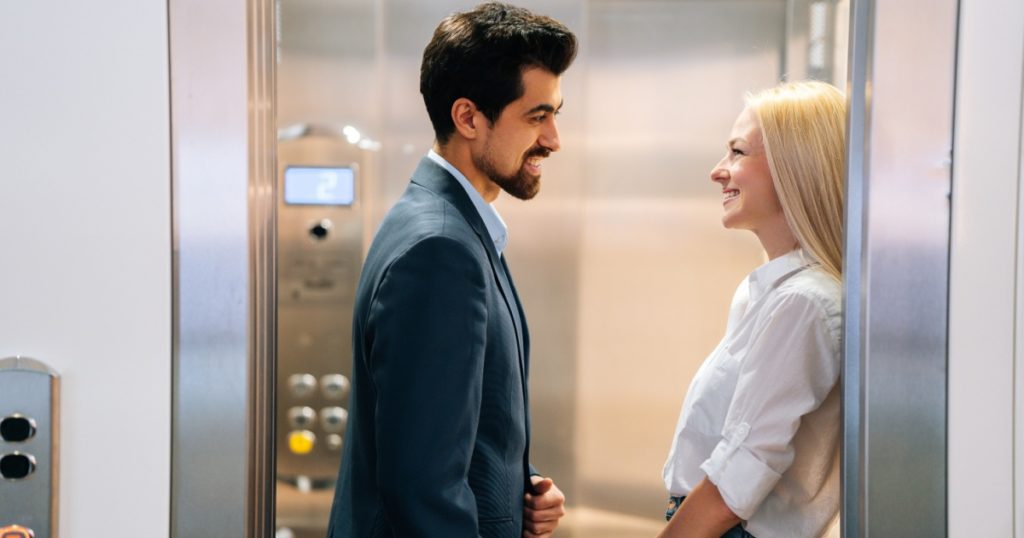 Handsome young man and beautiful woman looking at each other smiling standing inside of elevator in shopping mall. Cheerful business couple meeting in elevator of modern office building.