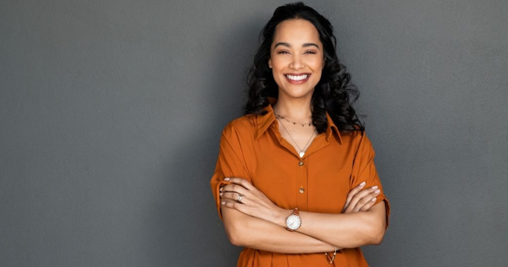 Portrait of a young latin woman with pleasant smile and crossed arms isolated on grey wall with copy space. Beautiful girl with folded arms looking at camera against grey wall. Cheerful hispanic woman