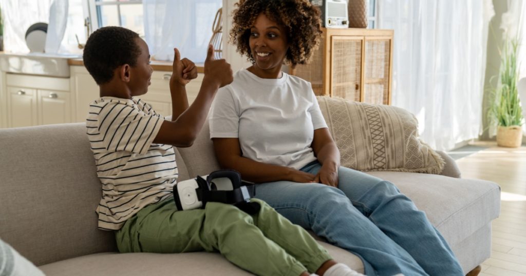 Joyful African American little boy making thumb up gesture for mother after play video game virtual reality headset. Pleased mom interested in opinion of son about VR 3D glasses for smartphone at home
