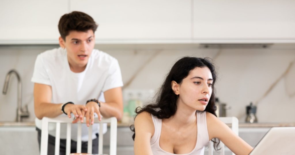 Sad and tired freelancer wife works at home on laptop and listens to her husbands reproaches in frustration. Family quarrel, lack of free time, unmet needs