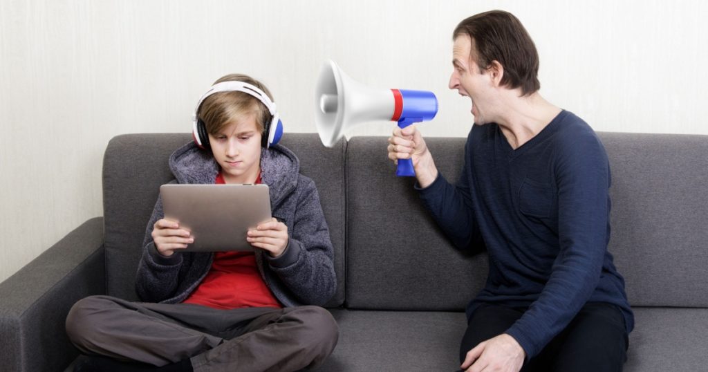 Tween son in headphones looks at the digital tablet display while his father yells at him through a megaphone