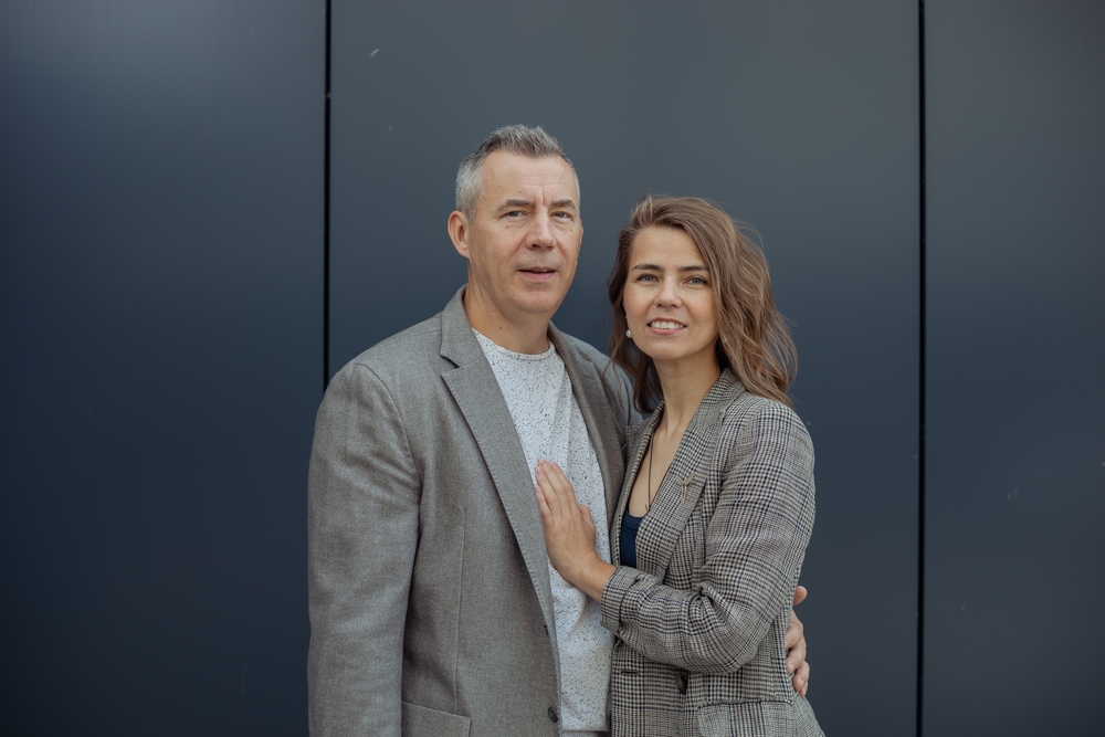 Romantic smiling couple of senior gray haired man and young blond woman embracing, look at camera against gray wall. Happy marriage couple. Age difference and generation gap in family. Copy space
