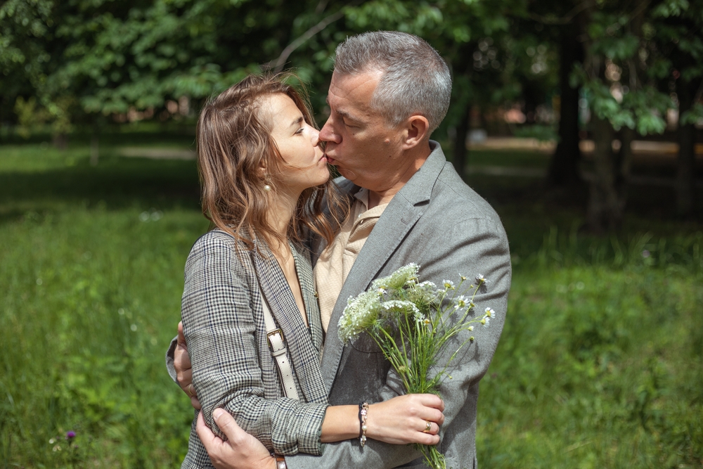 Romantic couple of two mature gray haired man and woman holding bouquet of flowers kissing and falling in love in green natural park outdoors. Happy marriage, loving close relationship in family.
