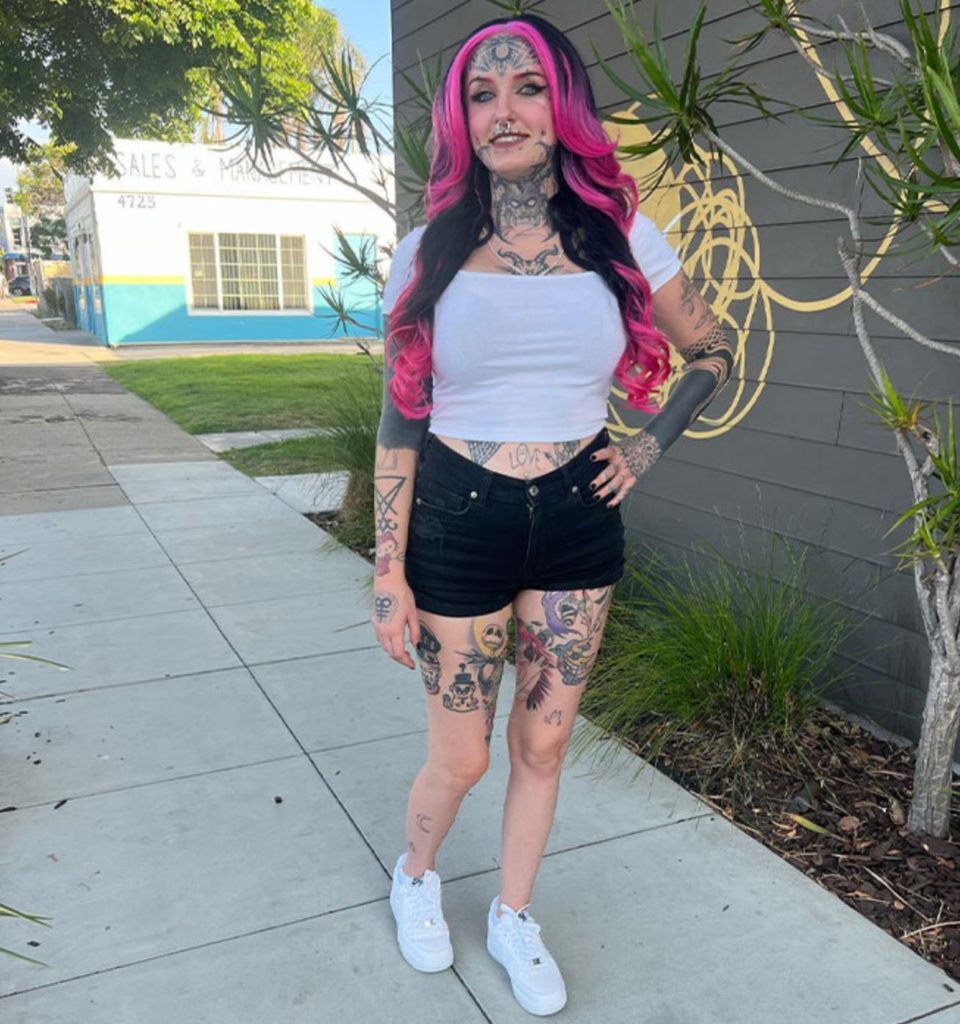 Woman with tattoos, piercings, and pink hair standing in front of a building with another building, trees, and a blue sky in the background. 
