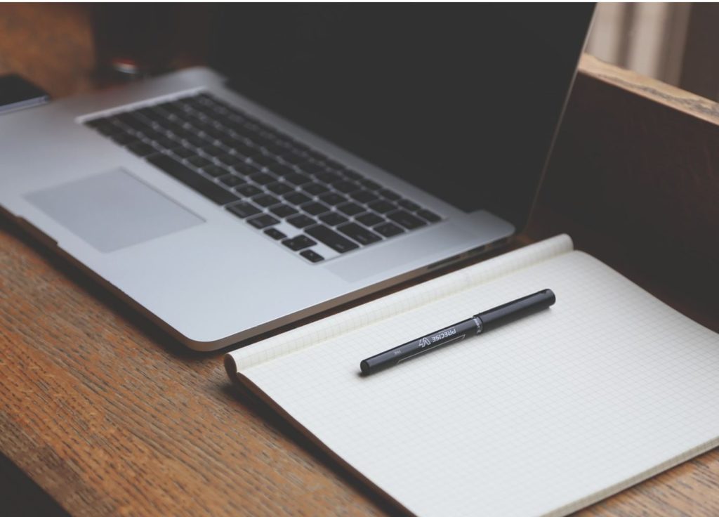 Empty notebook and pen next to a laptop on a wooden desk.