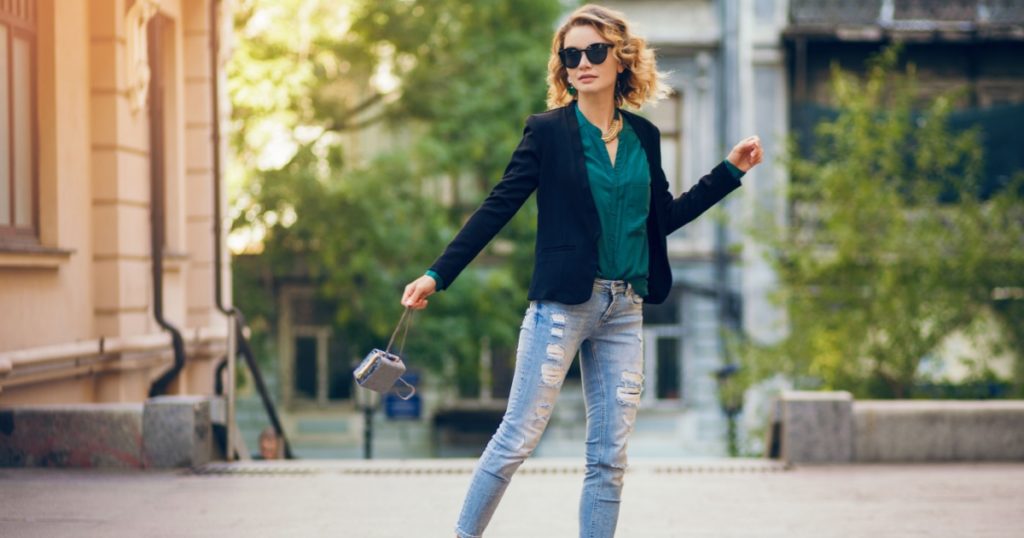 attractive elegant woman walking in city street on high heeled shoes, wearing blue jeans, black jacket, green blouse, sunglasses, holding little purse, fashion trend of summer, slim beautiful lady