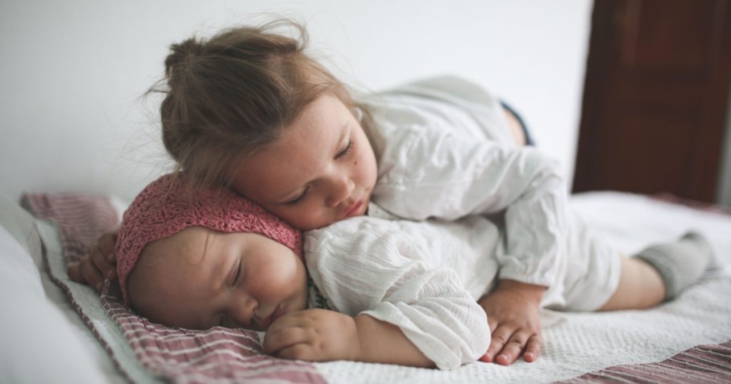 Toddler Girl Hugs a Sleeper Cute little baby in a crocheted cap on her stomach on a bed