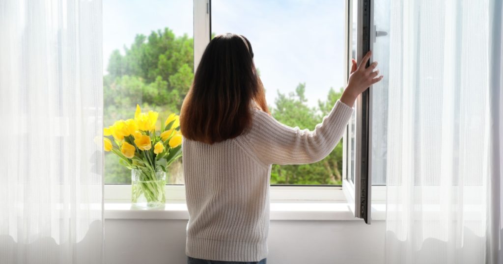 Pretty young Asian woman opening window at home on sunny day, back view
