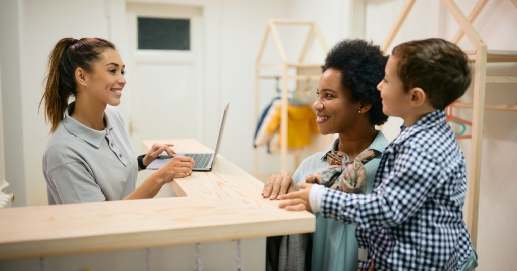 Happy kindergarten teacher communicating with African American mother and her son at reception desk.