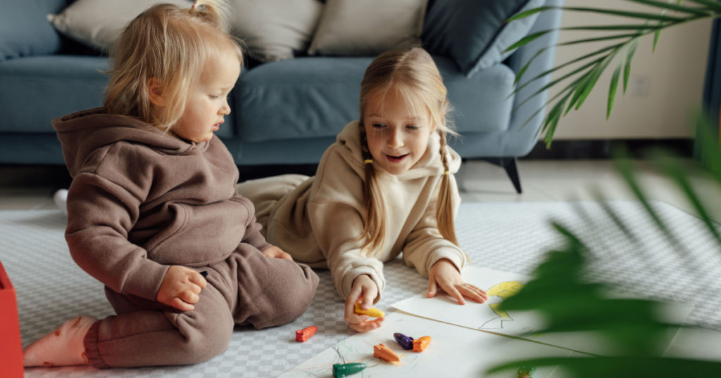 Little caucasian siblings drawing together on floor in living room at home. Happy girl eight years old and her baby sister painting with crayons. Art and crafts for toddler and preschooler