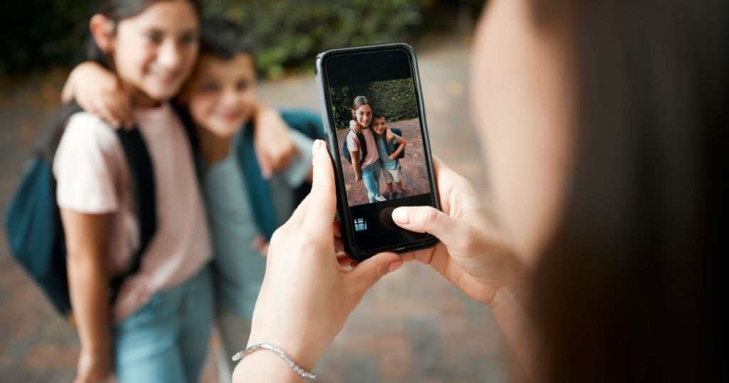 Children first day of school and mother taking photos with a phone of her cute kids. Closeup of screen picture of brother and sister embracing while posing for their mom outside in the morning