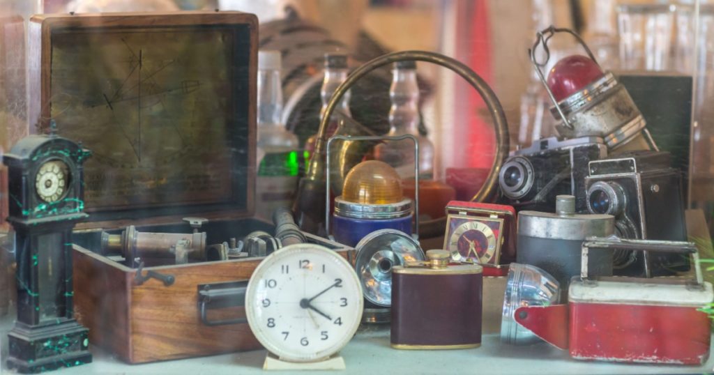 Assorted vintage items, clocks, cameras, flasks, sextant, lamps behind shop window.