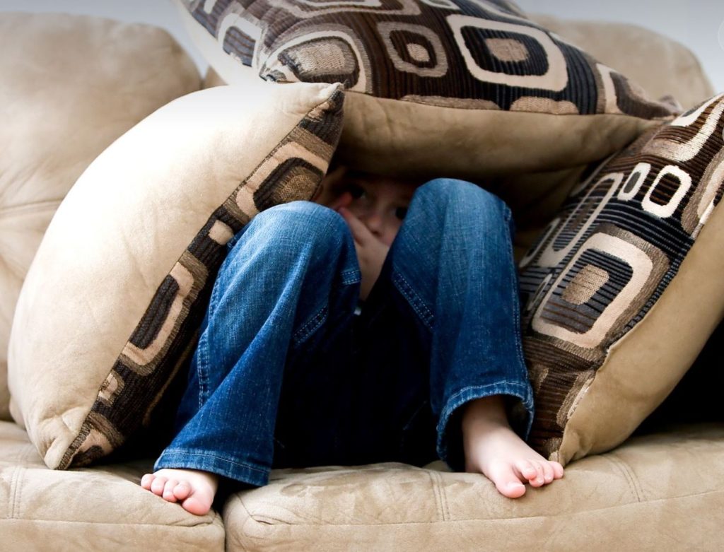 Person sitting on a tan couch and hiding in pillows. 