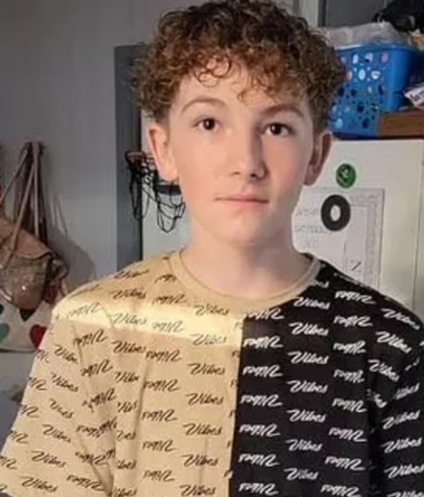 Boy wearing black and white shirt that says vibes in cursive. Purse, fridge, and laundry basket in the background.
