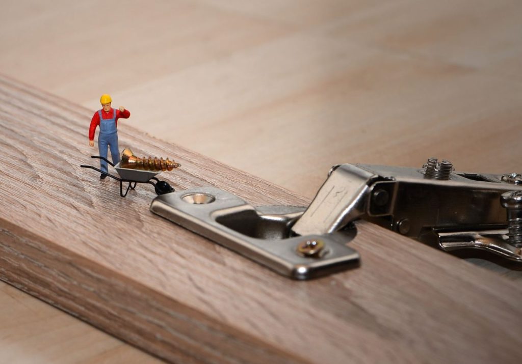 Tiny construction person with a wheelbarrow and screw. A door lock mechanism, and wood in the background. 